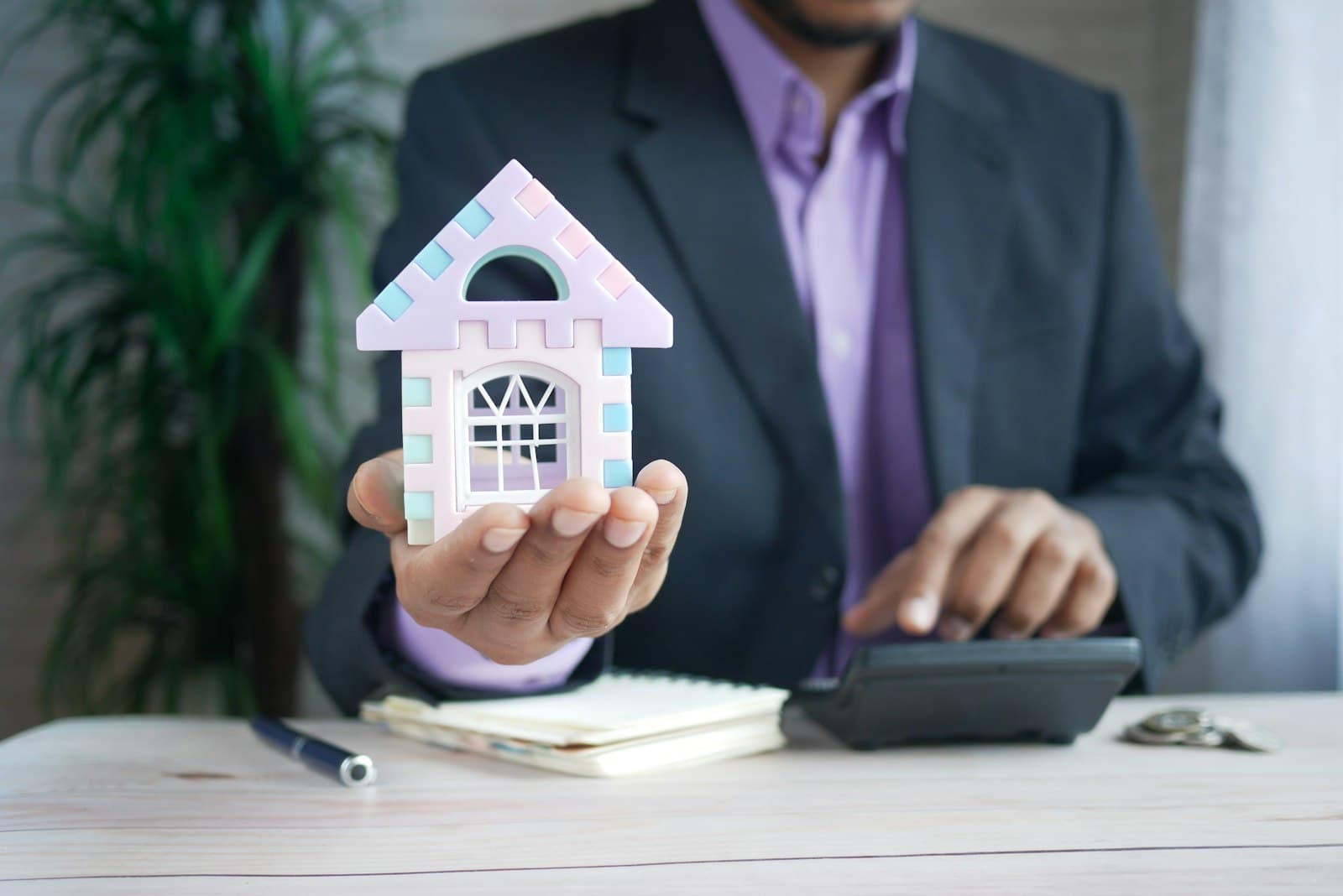 Person holding a small model house beside a calculator